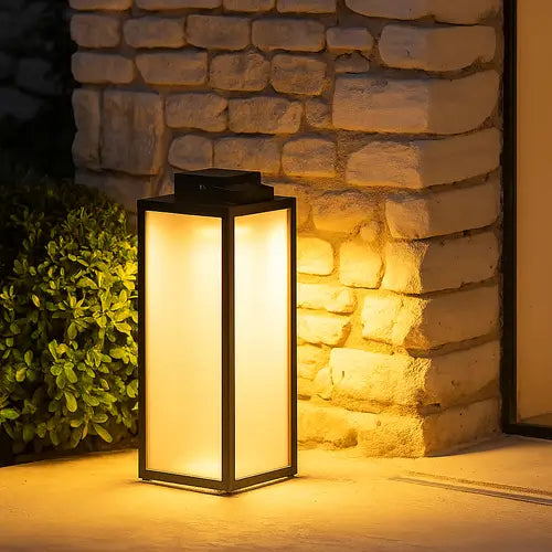 Lanterne LED extérieure minimaliste sur pied, forme rectangulaire avec cadre noir et panneau diffuseur opaque (givré), émettant une lumière chaude (jaune) et uniforme. La lanterne est posée au sol à l'entrée d'une maison devant un mur de pierres blanches/jaunes et un buisson. (Lanterne LED moderne, éclairage d'ambiance extérieur)