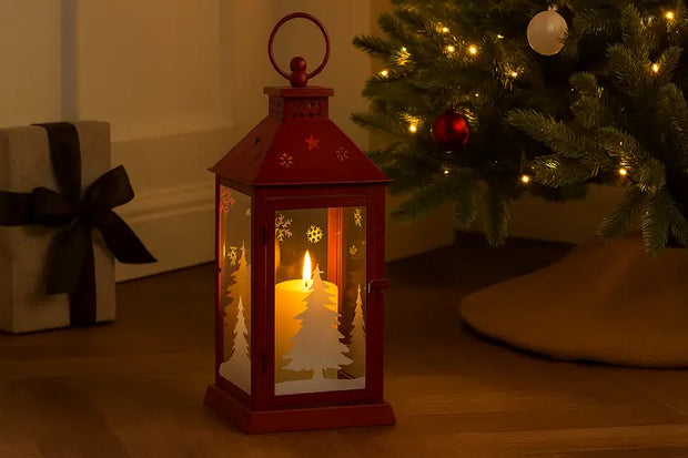 Lanterne de Noël rouge en métal avec découpes de flocons et de sapins de Noël, posée sur un plancher en bois à l'intérieur. Elle contient une grande bougie pilier allumée, créant une lumière chaleureuse. L'arrière-plan montre un arbre de Noël illuminé et un cadeau emballé. (Lanterne Noël rouge, décoration festive, sapin)