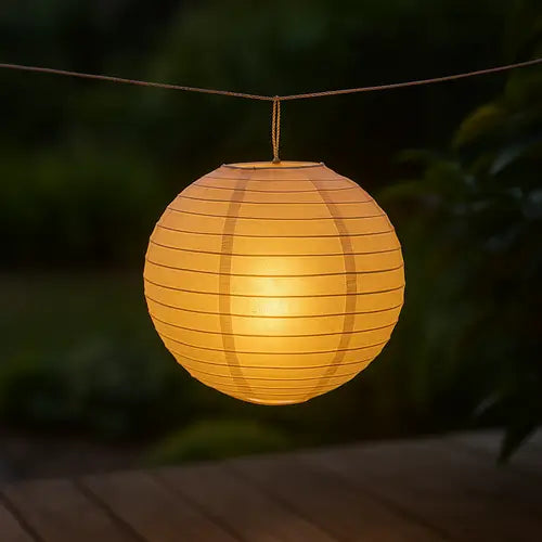 Lanterne ronde en papier de style japonais ou chinois, suspendue à une corde par une ficelle tressée en extérieur. La lanterne est éclairée de l'intérieur par une lumière chaude et dorée, créant une ambiance douce au crépuscule, au-dessus d'une terrasse en bois. (Lanterne papier suspendue, décoration jardin)