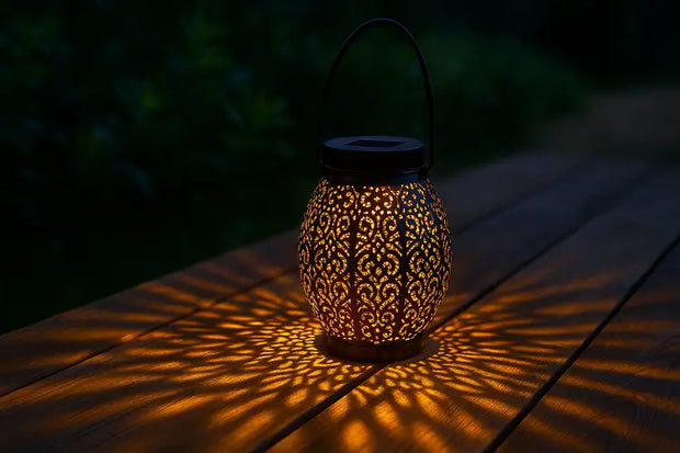 Lanterne solaire décorative en métal noir avec motifs découpés (orientaux ou floraux), posée sur une table en bois foncé en extérieur, projetant des ombres lumineuses dorées et complexes sur la surface. Le soleil est couché, créant une ambiance chaleureuse et captivante dans le jardin. (Lanterne solaire jardin, éclairage extérieur)